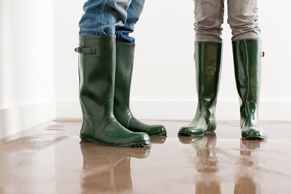 Why Is My Furnace Leaking Water? Image shoes two pairs of green rain boots standing on flooded floor inside home.